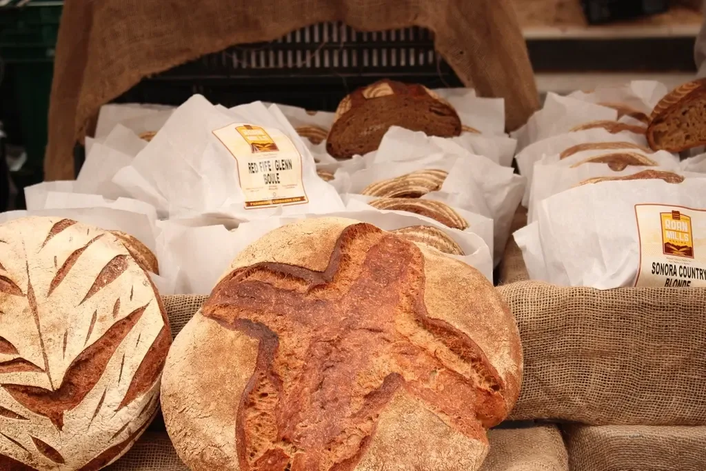 Runde Sauerteigbrote auf einem Markttisch, dahinter eingeschlagene Laibe in Papier